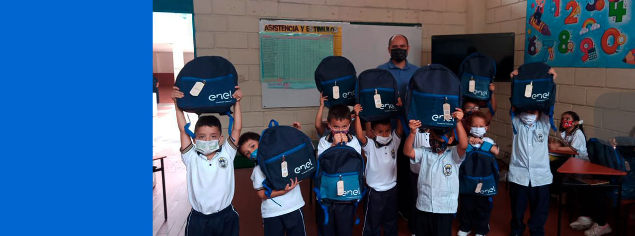 Grupo de niños pequeños, felices, en un salón con sudadera del colegio alzando maletas donadas por la empresa Enel