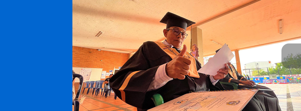 Foto de hombre de 58 años con traje de graduación y el diploma levantando el pulgar en señal de alegría