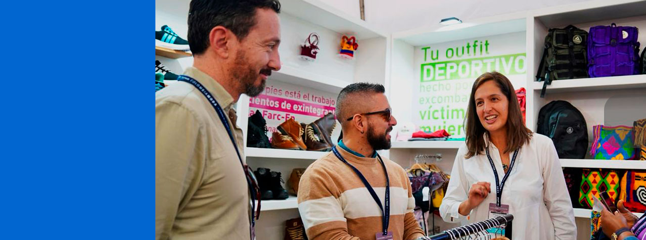 Foto del Stand de Los excombatientes de las FARC donde se ve de fondo estantes con zapatos y bolsos y están 3 personas hablando,