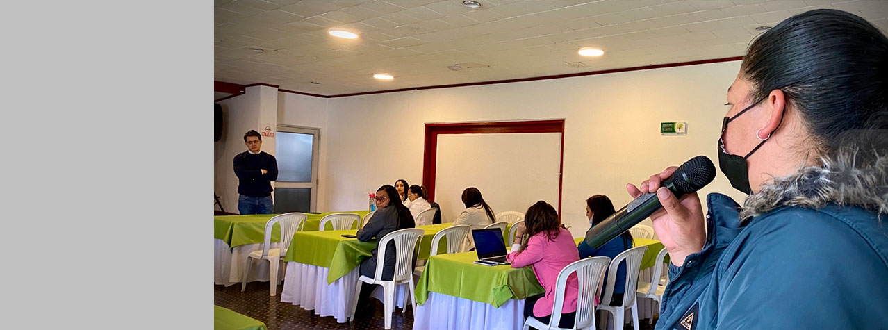 Grupo de personas en un salón sentadas en sillas y una persona está hablando por un micrófono 