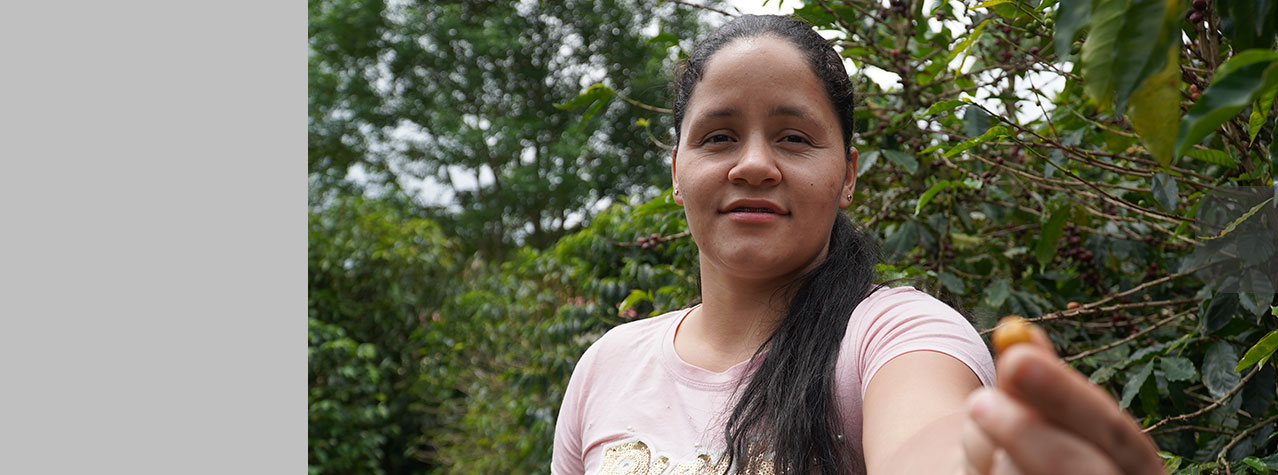 Foto de una mujer que mira de dfrente y tiene su mano estirada mostrando un grano de café