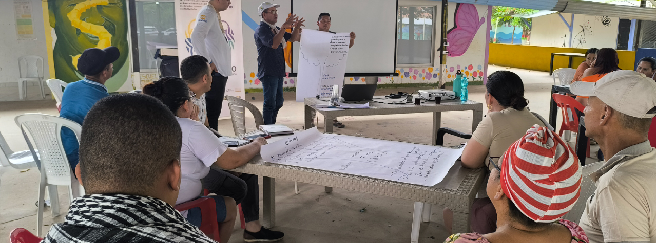 Grupo de personas reunidas, dos personas están exponiendo una cartelera. Otros poniendo atención.