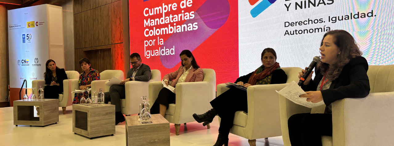 Grupo de personas sentados en sillas, en un escenario. Una mujer con un micrófono, hablando