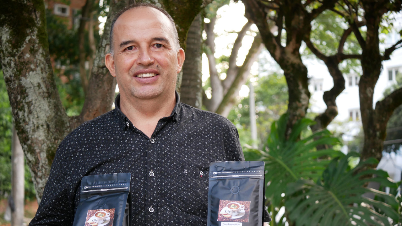 Persona sostiene dos paquetes de café Oh La La en un espacio exterior rodeado de árboles y vegetación.