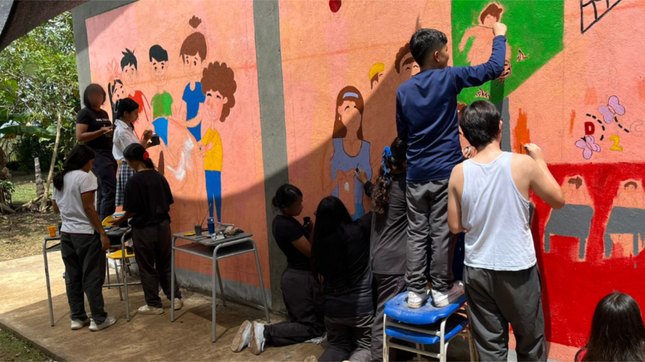Grupo de jóvenes pintando un mural colorido en una pared, representando escenas de infancia y aprendizaje