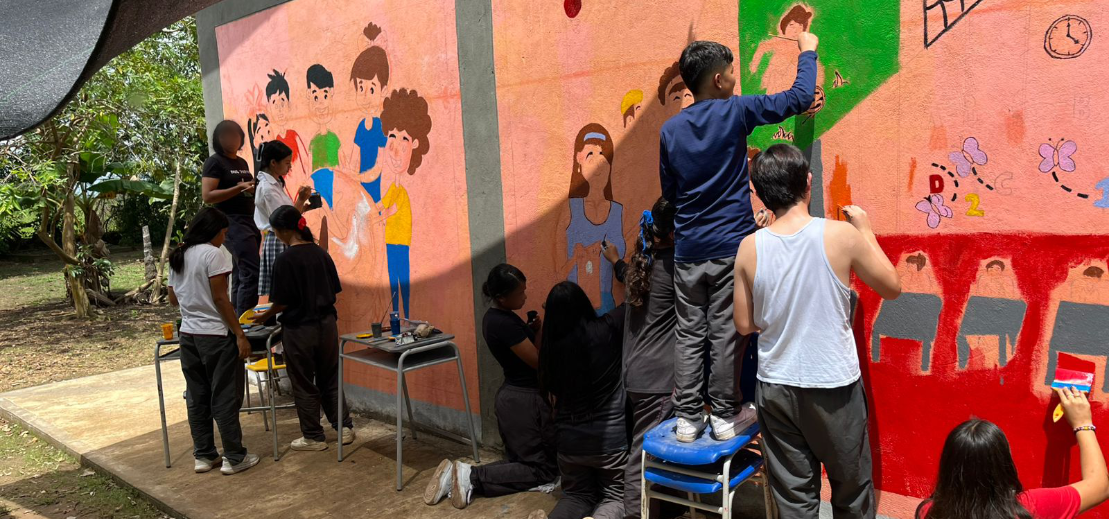 Grupo de jóvenes pintando un mural colorido en una pared, representando escenas de infancia y aprendizaje